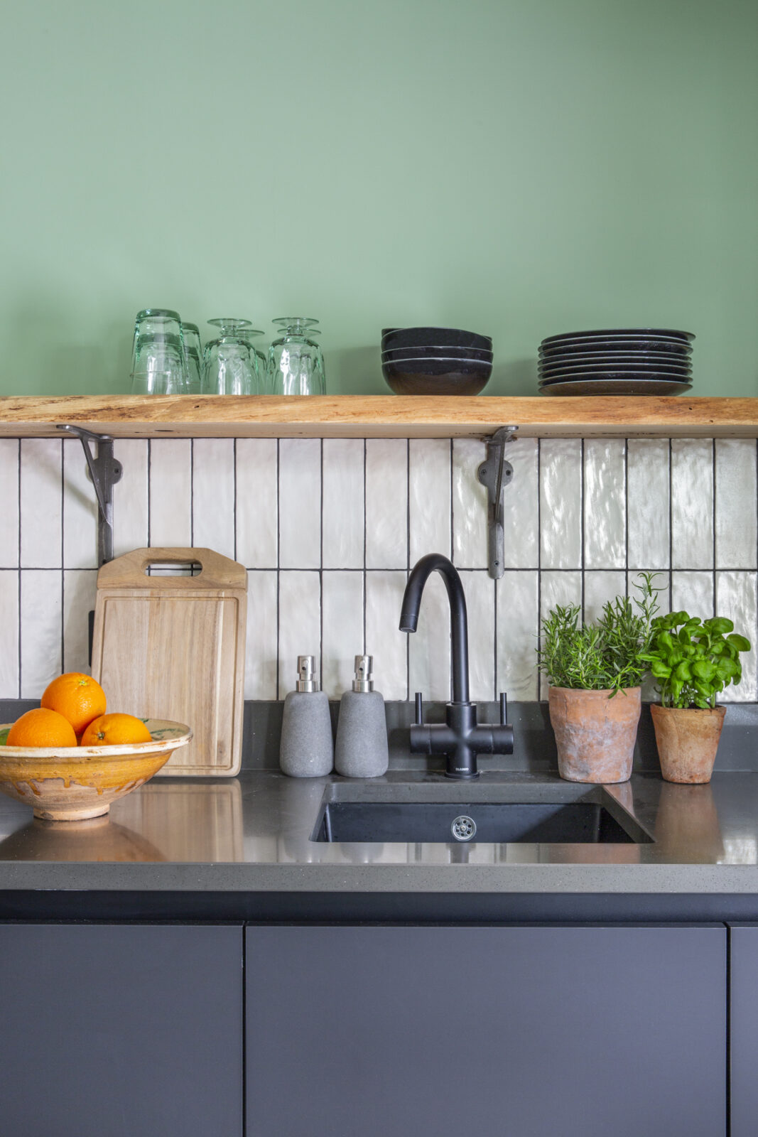 Close up of kitchen sink with charcoal units, exposed shelves, white tiles, green walls