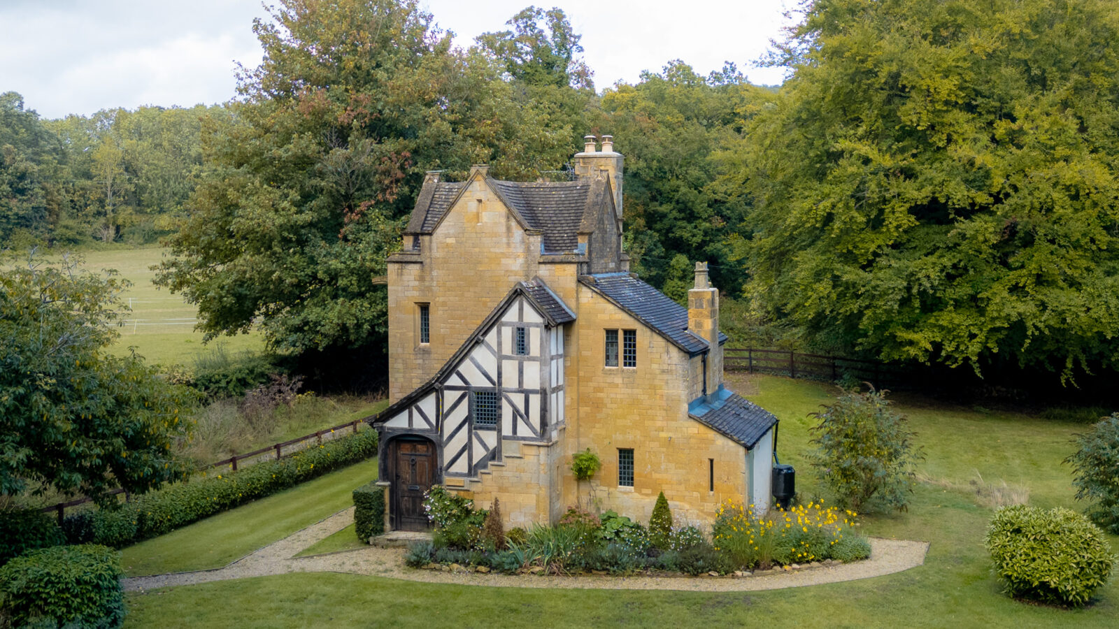 An aerial shot of a cotswold stone lodge with a Tudor style front in the middle of a secluded clearing with leafy trees and fields in the distance.